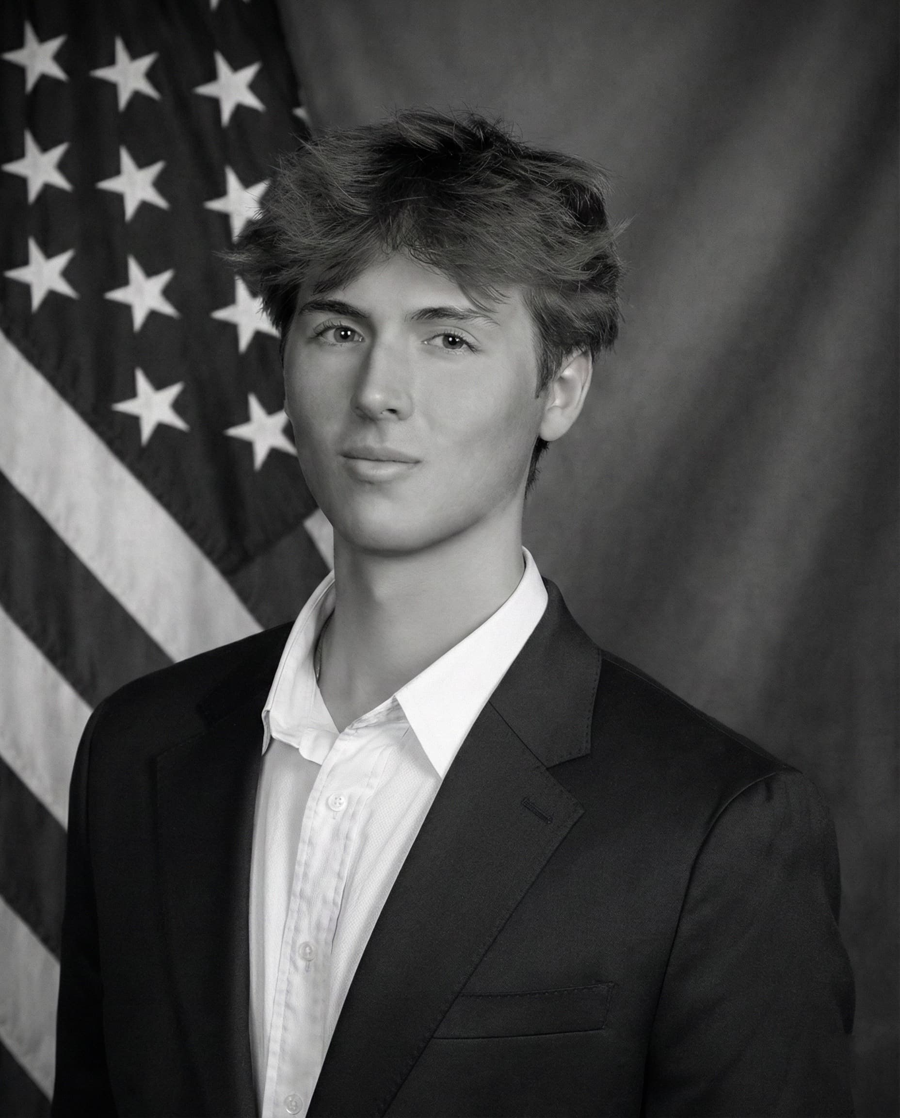 Black-and-white studio portrait of Caden Tacoronte in a dark suit and white shirt, photographed against a flag and drape.