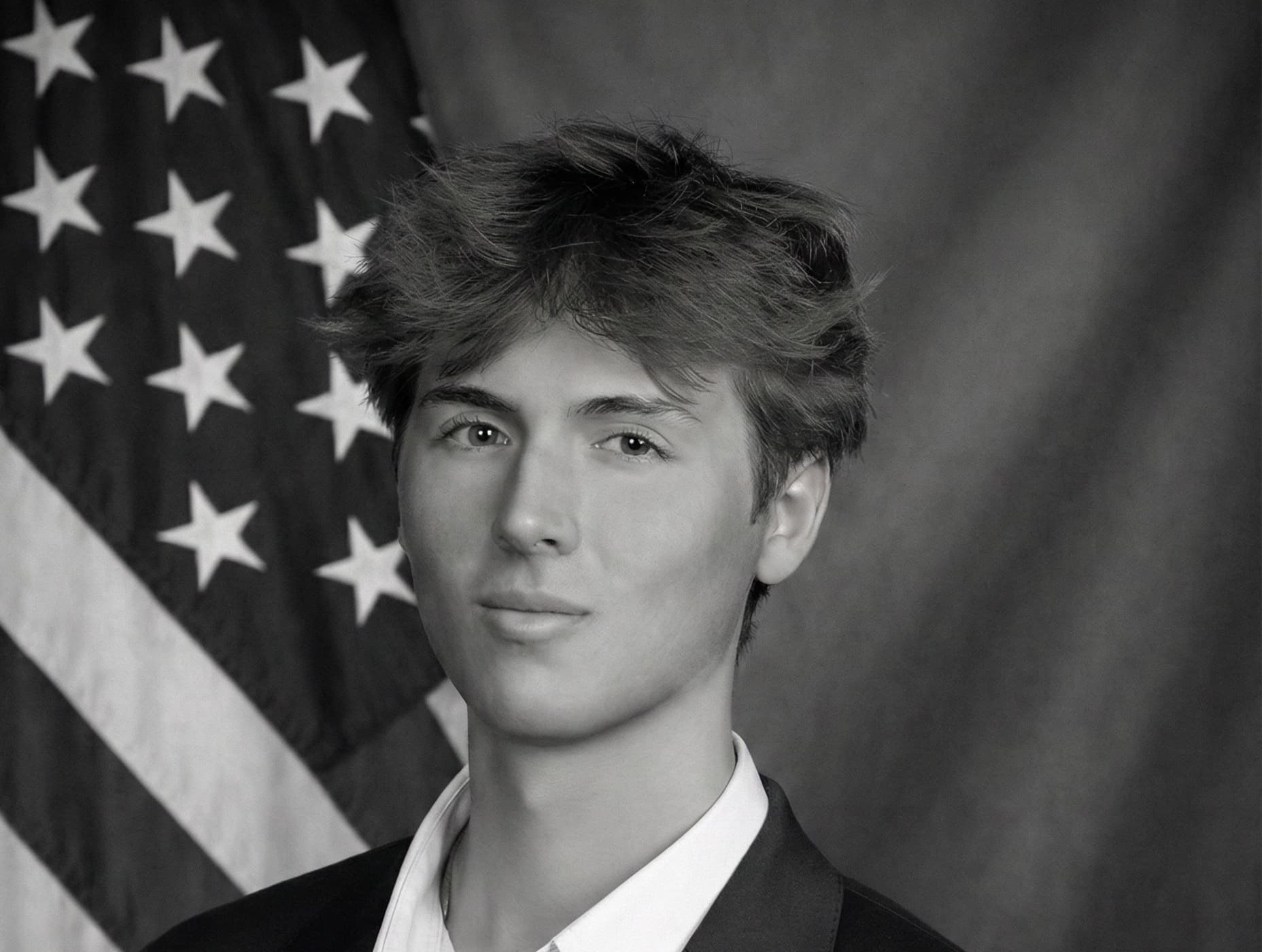 Black-and-white studio portrait of Caden Tacoronte in a dark suit, photographed against a flag and drape.