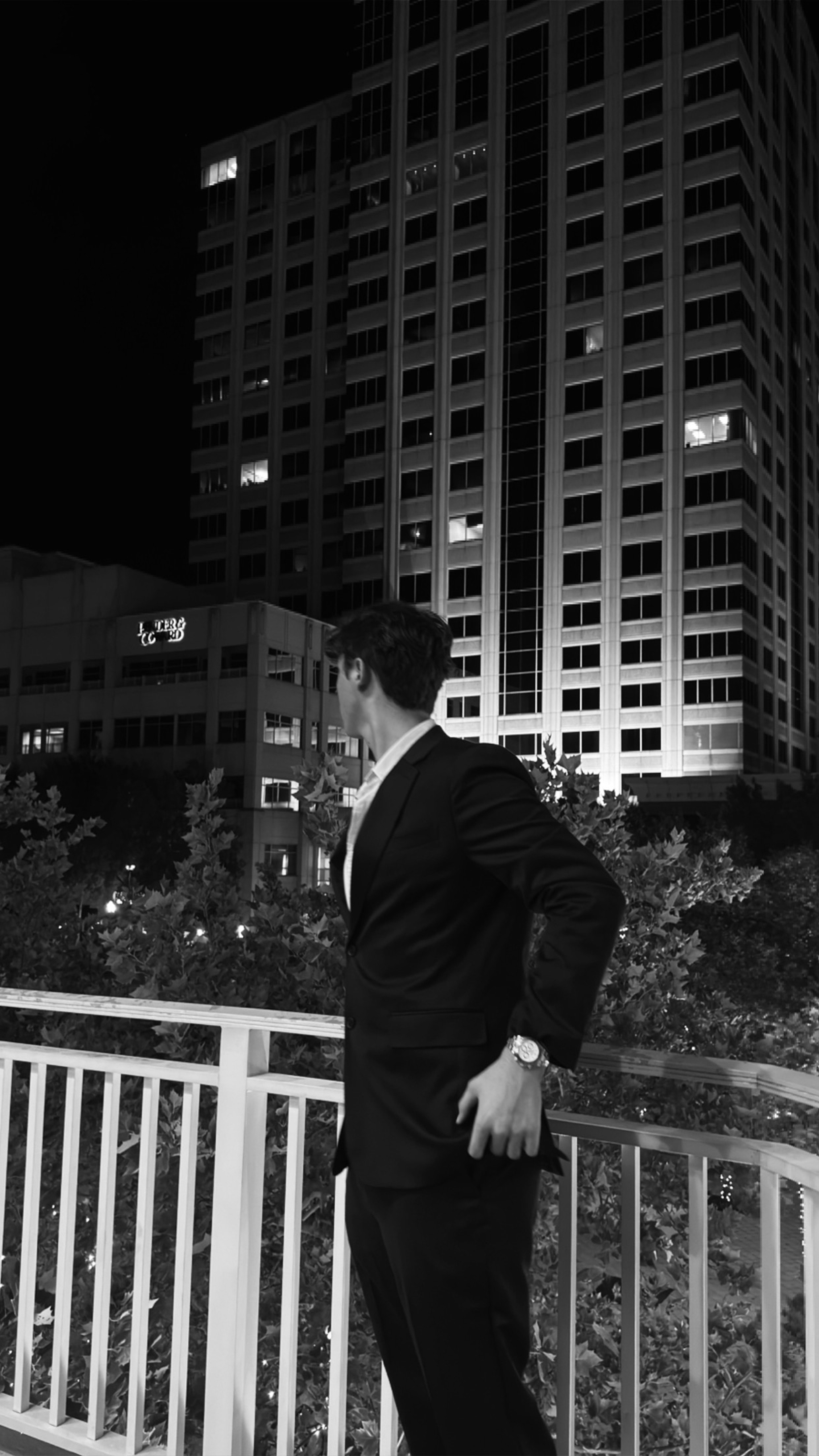 Caden Tacoronte in a dark suit at night, looking out over a downtown skyline from a terrace.